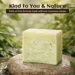 A rectangular bar of pale green soap standing upright on a wet stone, with blurred green foliage in the background. Text at the top reads, 'Kind to You & Nature' and 'Palm oil free formula made with eco-conscious values.