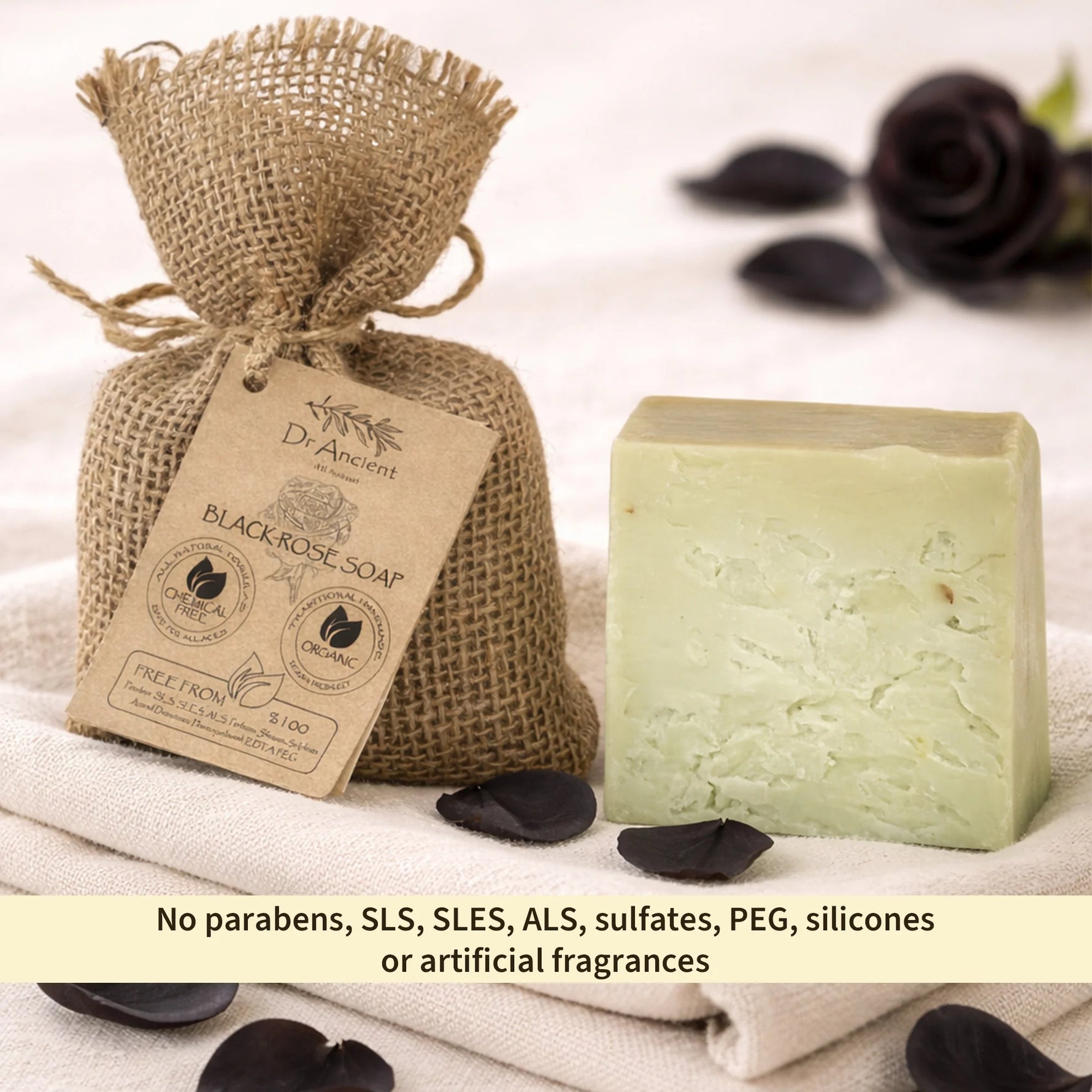 A bar of pale green soap stands beside a burlap drawstring pouch labeled Dr. Amandi Black Rose Soap, styled with dark rose petals on a soft beige fabric background.