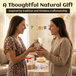 Two women smiling and exchanging a wrapped gift in a decorated birthday living room. Large text at the top reads 'A Thoughtful Natural Gift' with a subtitle 'Inspired by tradition and timeless craftsmanship.
