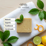 A rectangular bar of natural soap in a warm beige tone sits on a white textured towel, surrounded by green leaves, citrus peel, lime halves, and lemons on a light wood surface. The brand name 'DR.