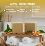 Two rectangular bars of pale yellow soap displayed on a small wooden stand in a spa-like setting, with white towels underneath and decorative leaves and orange peel around them. Text at the top reads 'Glow From Nature' and 'Nourishment & Purification In Every Bar' with bullet points about brightening complexion, boosting radiance, and smoothing texture.