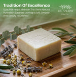 A rectangular bar of pale beige natural soap sits on a wooden board surrounded by eucalyptus leaves, dried lavender, and small yellow flowers. Text at the top reads 'Tradition Of Excellence' and 'GOAT MILK HELPS MAINTAIN THE SKIN'S NATURAL HYDRATION BALANCE, LEAVING IT SOFT, Smooth, And Deeply Nourished,' with the DR.