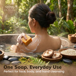 A woman sits in a hot spring-style bath holding a rectangular yellow cleansing soap bar, with white foam on her shoulder. The scene is outdoors in a lush green natural setting with bowls of ingredients and the text 'One Soap.