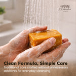 A person's hands covered in soap lather hold a rectangular bar of golden-yellow Dr. Ancient herbal cleansing soap under running water.