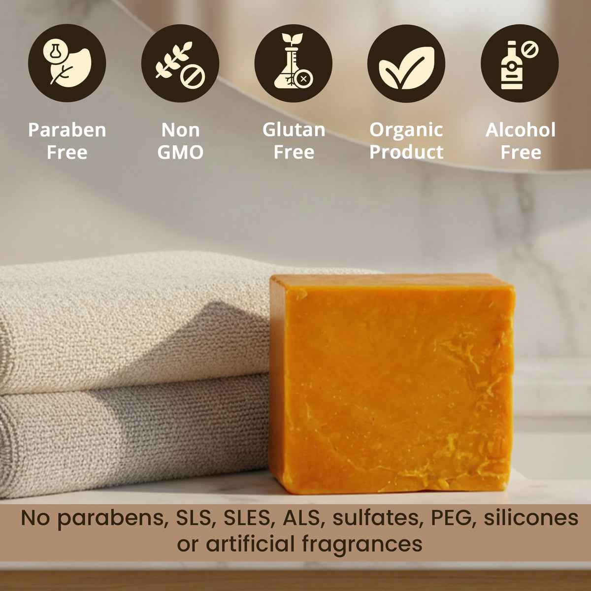 A rectangular bar of orange-yellow handmade soap stands in front of folded beige towels on a light marble surface. Five dark circular icons at the top read 'Paraben Free', 'Non GMO', 'Gluten Free', 'Organic Product', and 'Alcohol Free', with a text banner at the bottom stating no parabens, SLS, SLES, ALS, sulfates, PEG, silicones, or artificial fragrances.