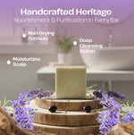 A handcrafted rectangular bar of pale yellow-green soap stands on stacked wood slices, surrounded by purple lavender flowers and small dark beads. The background is a softly blurred bathroom setting with white text overlays reading 'Handcrafted Heritage' and feature callouts including 'Non-Drying Formula,' 'Moisturizes Scalp,' and 'Deep Cleansing Action.