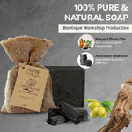A bar of charcoal soap in a burlap bag with a paper label, shown alongside loose charcoal pieces and green olives on a gray textured background. Text reads '100% Pure & Natural Soap' and 'Boutique Workshop Production,' with icons for natural plant oils and activated charcoal.