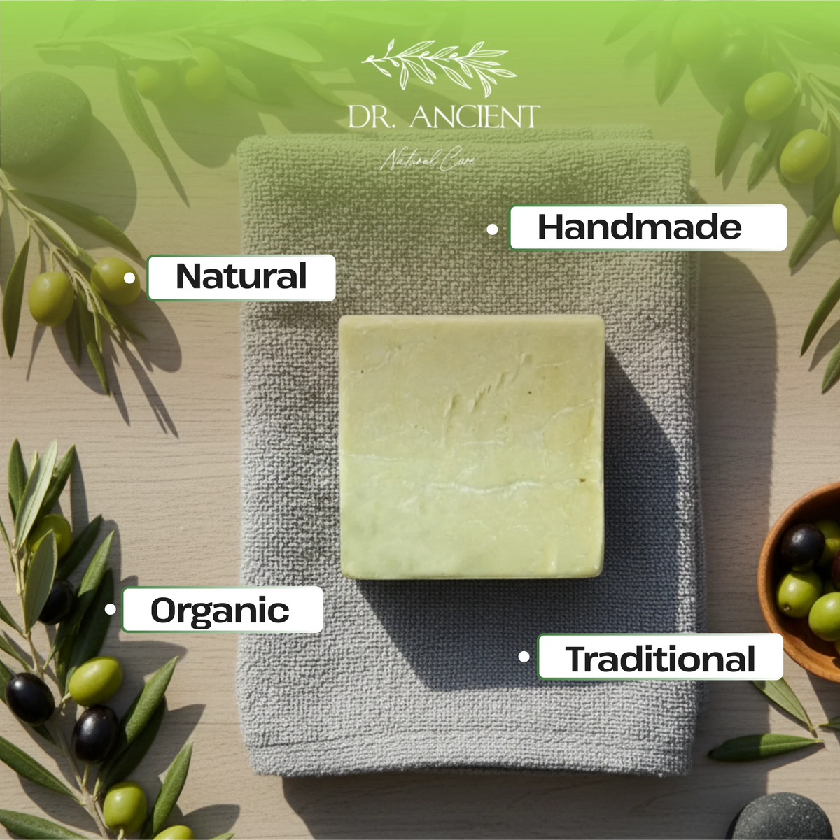 A rectangular bar of pale yellow-green soap rests on a folded gray textured towel, surrounded by olive branches and olives on a light wood surface. White labels point to the soap with the words Natural, Handmade, Organic, and Traditional.