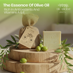 A bar of pale yellow-green soap stands beside a small burlap-wrapped gift bag with a paper tag, styled on two round wooden platters with olive branches and green olives. Text at the top reads 'The Essence Of Olive Oil' and 'Rich In Antioxidants And Vitamins A & E' with the brand 'DR.
