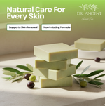 Four pale yellow-green bars of soap stacked on a light stone surface with olive branches and olives around them. Large text at the top reads 'Natural Care For Every Skin' with the Dr.