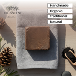 A rectangular bar of brown soap rests on a folded light gray cloth on a light wood surface, surrounded by pinecones, greenery, a small scoop of dark powder, and text labels reading Handmade, Organic, Traditional, and Natural. The Dr.