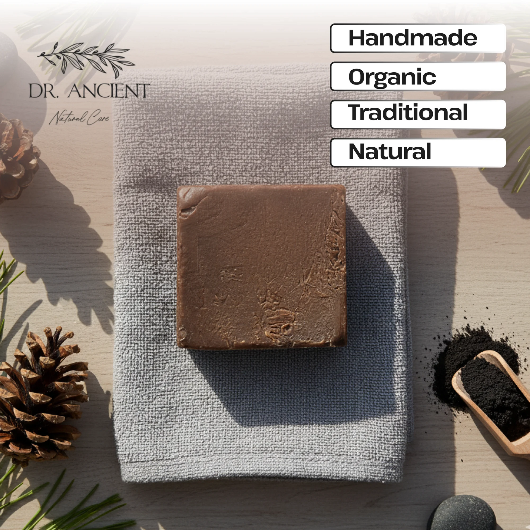 A rectangular bar of brown soap rests on a folded light gray cloth on a light wood surface, surrounded by pinecones, greenery, a small scoop of dark powder, and text labels reading Handmade, Organic, Traditional, and Natural. The Dr.