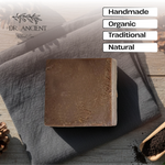 A rectangular bar of brown soap rests on a dark gray cloth over a rustic wooden surface, with pinecones and a small wooden scoop of dark powder nearby. White text boxes on the right read 'Handmade', 'Organic', 'Traditional', and 'Natural', and the brand name 'DR.