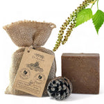 A bar of Dr. Ancient Pine Tar Soap in a rustic brown color, displayed next to its burlap packaging bag with a kraft paper tag, a pinecone, and green birch leaves on a white background.