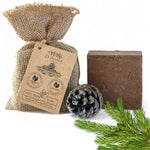 A bar of brown soap stands beside a burlap drawstring bag with a kraft paper tag, a pinecone, and green pine branches on a white background. The tag reads Dr.