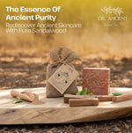 A bar of Dr. Ancient Natural Care sandalwood skincare soap stands on a wooden board outdoors, alongside a small burlap drawstring pouch with a paper tag, a wooden scoop of powder, and a few green leaves.