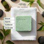 A rectangular bar of pale mint green soap rests on a folded white towel, surrounded by natural props including green leaves, dark stones, and a wooden scoop of green powder. The brand name 'DR.