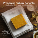 A rectangular bar of golden yellow handmade soap rests on a folded white linen cloth on a dark wooden surface. The scene includes a small glass bottle of oil, dried flowers, a bowl, and the text 'Preserves Natural Benefits' and 'DR.
