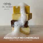 A stack of five natural soap bars in pale yellow, cream, and dark brown sits on a marble surface with white foam dripping down the sides. The brand logo 'DR.