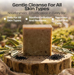 A rectangular bar of beige herbal soap stands on a wooden stump outdoors, surrounded by olive leaves and small black seeds. Text at the top reads 'Gentle Cleanse For All Skin Types' with bullet points about nourishment, purification, safety, soothing irritation, and removal of dead skin cells.