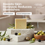 A bar of pale yellow soap stands on stacked stone blocks with olive branches, olives, and a small bowl of olives in a softly lit bathroom-like setting. Text on the image reads 'Boosts Skin Renewal, Reduces Blemishes' and includes the brand 'DR.