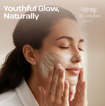 A woman with closed eyes applies a white facial cleanser or mask cream to her face, with soft warm lighting and the text 'Youthful Glow, Naturally' and the brand 'DR. ANCIENT Natural Care' in the upper right.