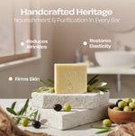 A bar of pale yellow soap stands on stacked stone blocks with olive branches and olives in a softly lit bathroom setting. Text at the top reads 'Handcrafted Heritage' with additional marketing copy about reducing wrinkles, restoring elasticity, and firming skin.