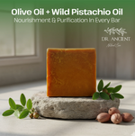 A rectangular bar of orange-yellow soap stands on a rough stone slab with olive leaves and pistachio nuts around it. Large text at the top reads 'Olive Oil + Wild Pistachio Oil' and 'Nourishment & Purification In Every Bar,' with the brand 'DR.