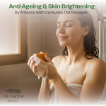 A woman wrapped in a white towel stands in a warm bathroom setting, holding a bar of soap with lathered hands. Large text at the top reads 'Anti-Ageing & Skin Brightening By Artisans With Centuries Old Receipts,' and the brand name 'DR.