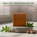 An orange-brown rectangular bar of soap stands upright on a rough stone slab, surrounded by green leaves and whole pistachio nuts. Text at the top reads 'Olive Oil + Wild Pistachio Oil' and 'Nourishment & Purification In Every Bar,' with the Dr.