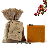 A bar of orange-yellow herbal skin soap next to a burlap pouch with a paper tag reading Dr. Aumont BYLINE SOAP, with small dark berries scattered on a white background and pink flowers in the corner.