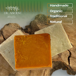 Two rectangular bars of soap displayed on a rustic wood slice background, with one bar in front in a warm amber-orange tone and the other behind in a pale creamy beige tone. The image includes the brand name Dr.