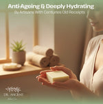 A woman in a beige robe holds a rectangular bar of pale yellow soap in her hands, with a softly lit spa-like interior in the background. Large text at the top reads 'Anti-Ageing & Deeply Hydrating By Artisans With Centuries Old Receipts,' and the brand 'DR.