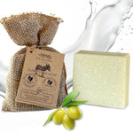 A bar of Dr. Ancient Donkey Milk Soap shown next to its burlap pouch packaging with a kraft paper tag, and green olives on a white background with a milk splash graphic.
