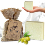 A bar of pale yellow-green donkey milk soap is shown in a hand, alongside another soap bar, a burlap-wrapped gift bag with a paper tag reading Dr. Ancient, and green olives on a white background with a splash graphic.