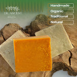 Three rectangular bars of natural soap displayed on a rustic wood background, with one orange bar in front and two pale beige bars behind. The image includes branding for Dr.