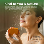 A woman with closed eyes holds an orange bar of soap in her hands, with water droplets falling around her face. Text at the top reads 'Kind To You & Nature' and 'Crafted With Olive & Turmeric Oils For Skin & Hair Wellness,' with 'DR.